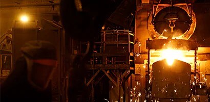 Fundición industrial con horno vertiendo metal incandescente. Trabajador con equipo de protección a la izquierda, rodeado de chispas y brillo del metal.
