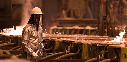Un trabajador con equipo de protección, incluyendo un casco y una chaqueta reflectante, supervisa el proceso de fundición en una planta industrial, rodeado de maquinaria y metal fundido.
