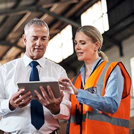 A man wearing a tie holds a tablet and a woman wearing a fluorescent safety vest points to the tablet.