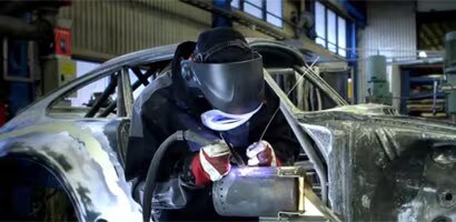 Un trabajador con equipo de protección, incluyendo casco y guantes, realiza labores de soldadura en el chasis de un automóvil dentro de un taller industrial.
