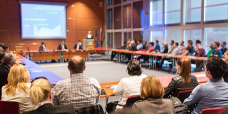 Very large gathering of people seated at a large square table viewing a graph being displayed.

