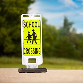 Standalone sign with yellow background and black image of two people crossing street and words “school crossing.”