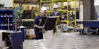Video still of workers at a machine on the assembly line
