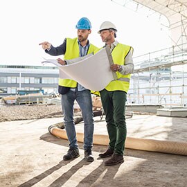 Two construction managers in hard hats, with one pointing and one holding construction blueprints.
