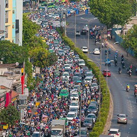 Urban road with numerous cars, motorcyclists and pedestrians crowding one side.