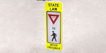 Standalone sign with symbols and letters to create the message “State law: Yield to pedestrians within crosswalk.”

