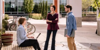 Two women and man standing around talking on a corporate plaza in the sun.
