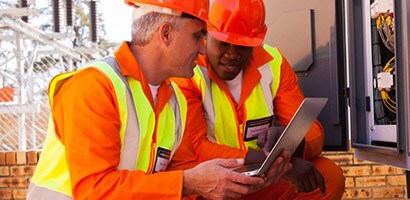 Dos hombres que visten de color naranja miran una computadora que muestra videos de capacitación de 3M relacionados con la instalación de productos eléctricos de 3M.
