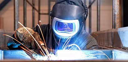 Un soldador con casco de protección trabaja en una estructura metálica, generando chispas y luz intensa en un entorno industrial.
