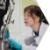 A woman in a white lab coat with safety glasses on watching an ongoing experiment.